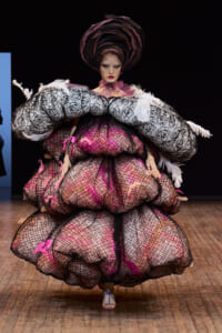 Model on a runway wearing an oversized pink and black mesh gown with rose-pattern sleeves and a dramatic circular headpiece.