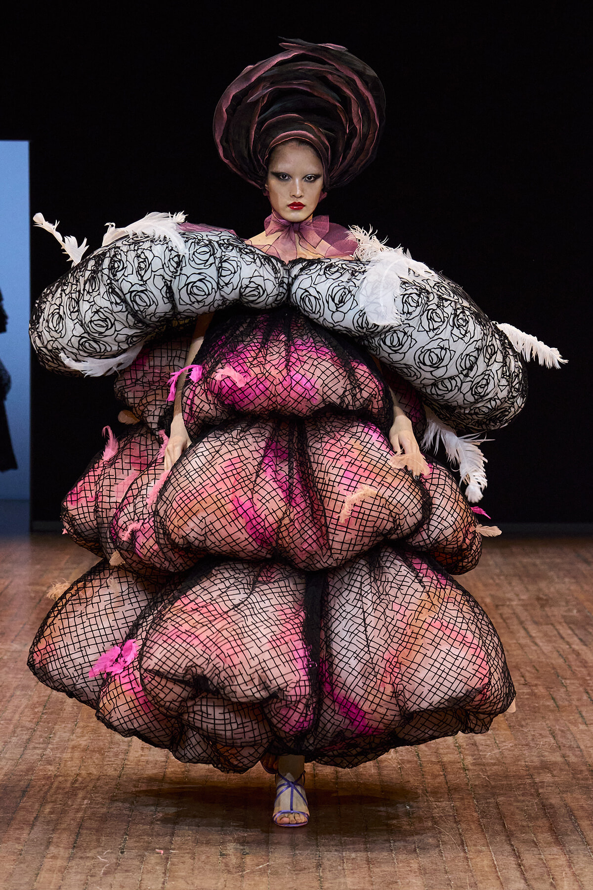 Model on a runway wearing an oversized pink and black mesh gown with rose-pattern sleeves and a dramatic circular headpiece.