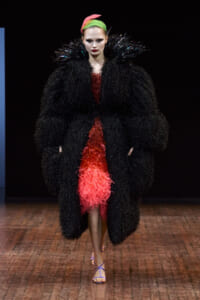 Runway model in a voluminous black feathered coat over a textured red dress, with a neon pink-green headband and purple sandals.