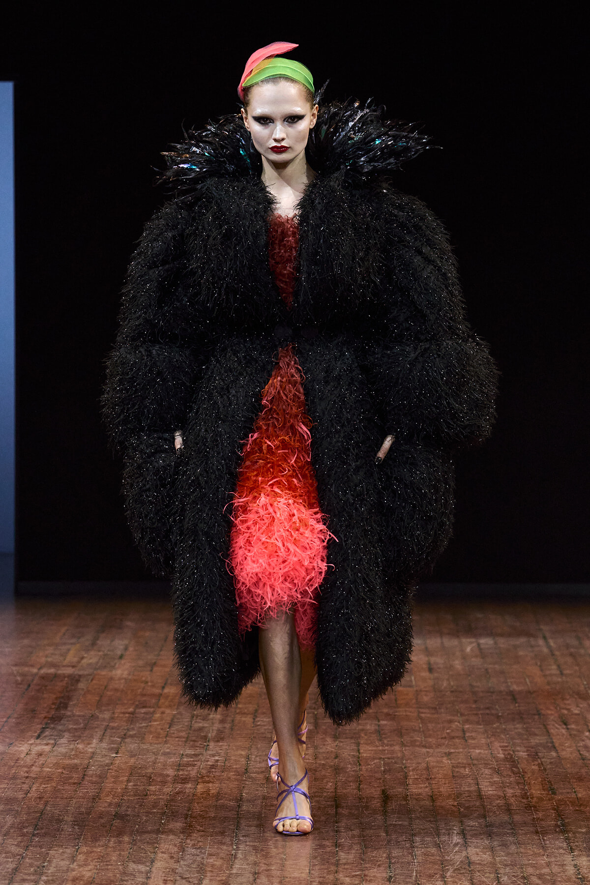 Runway model in a voluminous black feathered coat over a textured red dress, with a neon pink-green headband and purple sandals.