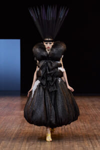 Runway look: black glittery couture gown with oversized sculpted shoulders and a tall feathered headpiece on a dark stage, model walking forward