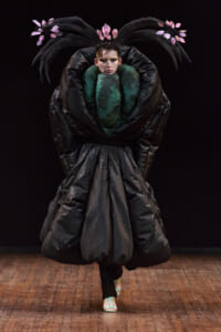 Model on a runway wearing a dramatic oversized black sculptural gown with a pink leaf headpiece and a green bodice centerpiece on a dark backdrop.
