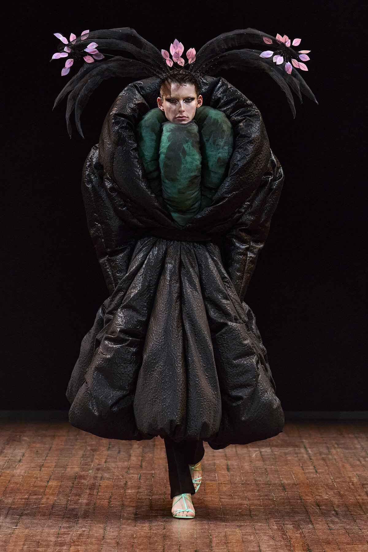 Model on a runway wearing a dramatic oversized black sculptural gown with a pink leaf headpiece and a green bodice centerpiece on a dark backdrop.