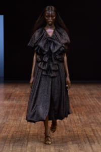 Model walks the runway in a metallic dark-gray, glittery gown with dramatic ruffled layers and a mid-length skirt, paired with strappy sandals