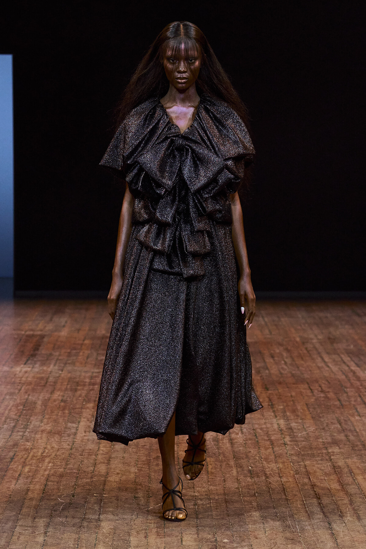 Model walks the runway in a metallic dark-gray, glittery gown with dramatic ruffled layers and a mid-length skirt, paired with strappy sandals