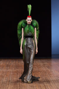 Model on a runway wearing a dramatic green sheer gown with large sculptural shoulders and a tall green headpiece.
