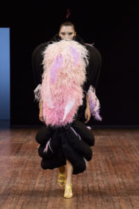 Model on runway wearing voluminous pink feathered cape over black outfit with yellow strappy heels