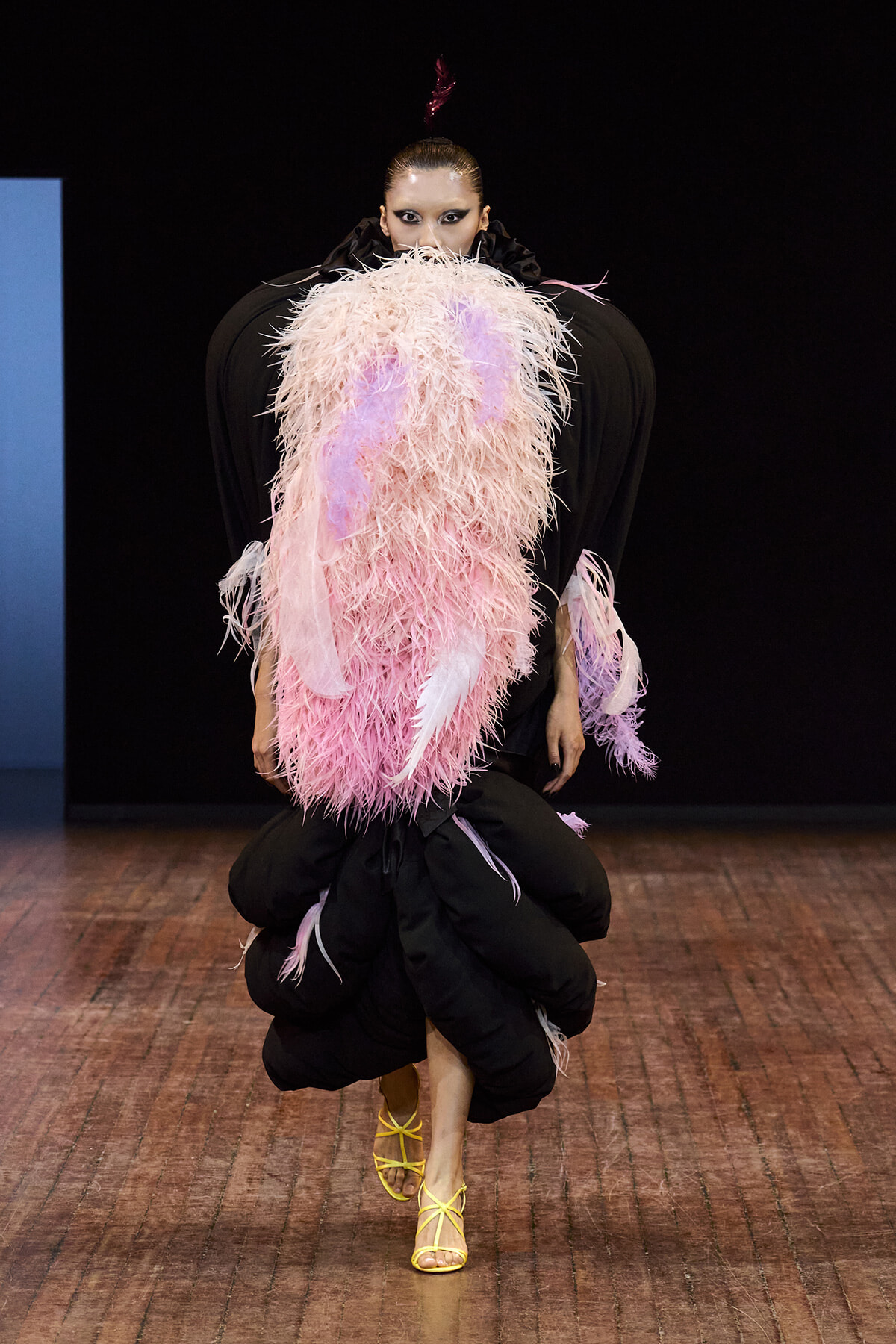 Model on runway wearing voluminous pink feathered cape over black outfit with yellow strappy heels