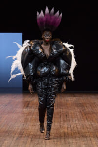 Model on a runway wearing a dramatic black glossy jumpsuit with oversized padded shoulders, white feather wings, and a tall purple feather headdress.
