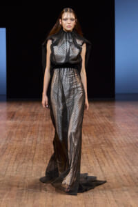 Model on a runway wearing a sheer black polka-dot gown with high neck and dramatic cape-like shoulders.