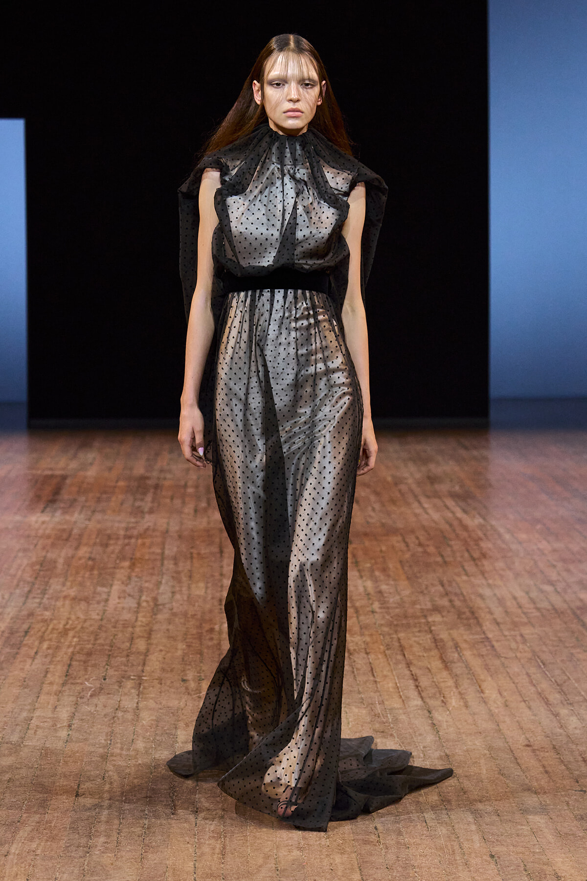 Model on a runway wearing a sheer black polka-dot gown with high neck and dramatic cape-like shoulders.