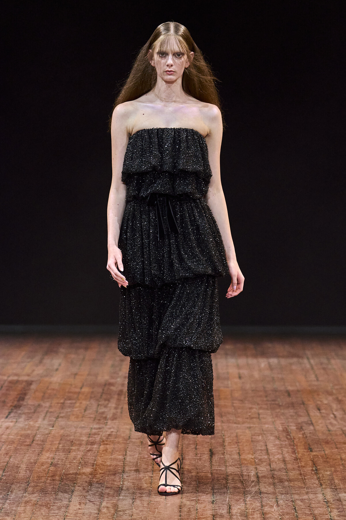 Model on the runway wearing a strapless, black glitter-tinted tiered dress with strappy sandals.
