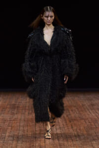 Model walking a fashion runway in a black, shaggy-textured coat with a deep V neckline and strappy sandals.