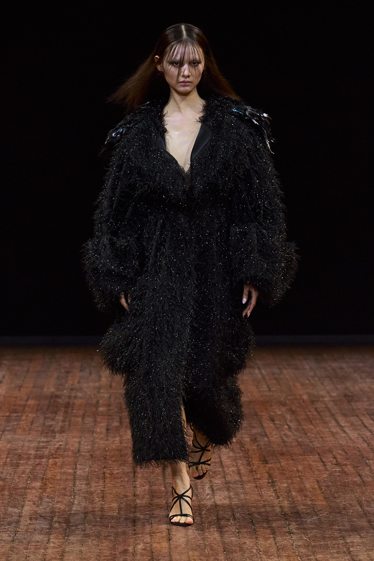 Model walking a fashion runway in a black, shaggy-textured coat with a deep V neckline and strappy sandals.