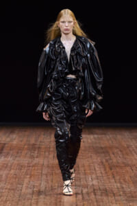 Model walking a fashion runway in a glossy black patent leather ensemble with oversized ruffled collar, voluminous sleeves, and ruched pants, against a dark background.
