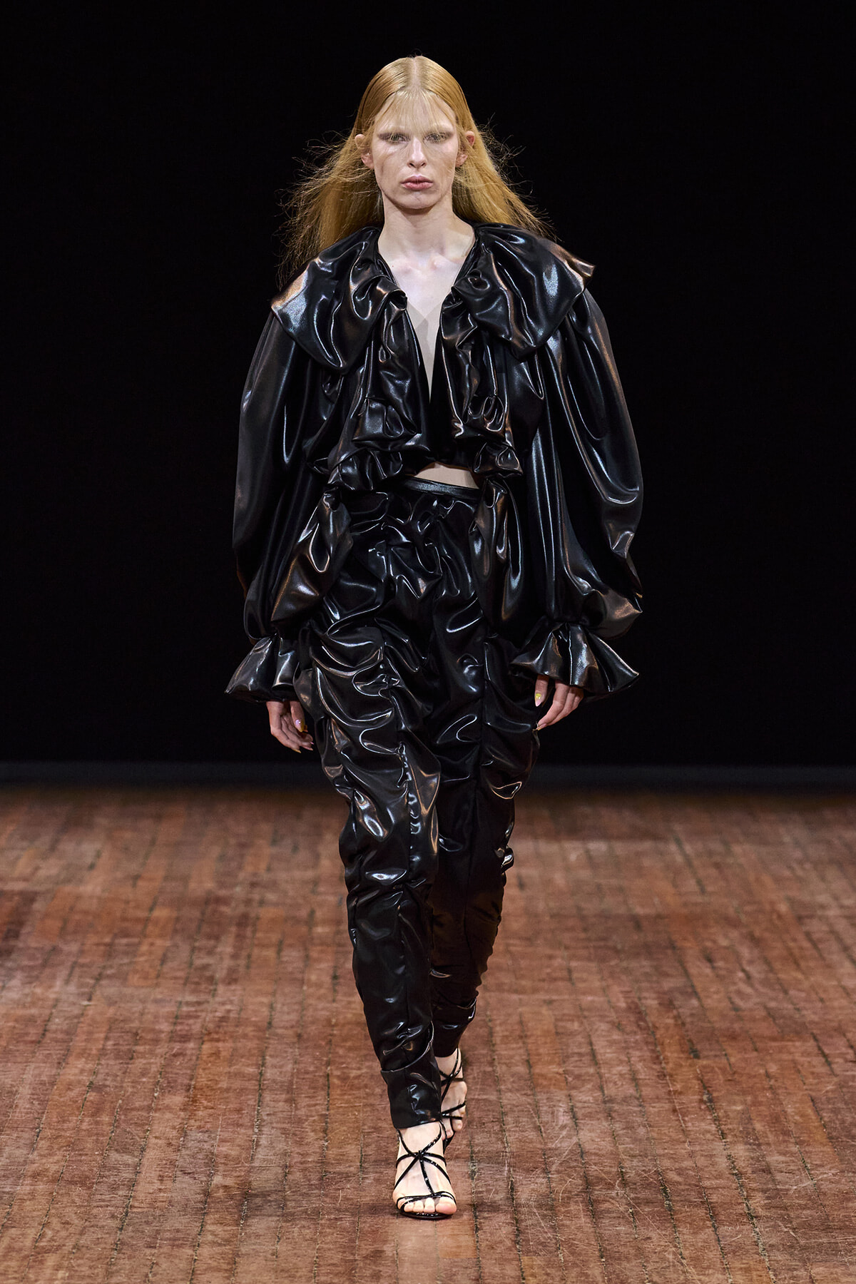 Model walking a fashion runway in a glossy black patent leather ensemble with oversized ruffled collar, voluminous sleeves, and ruched pants, against a dark background.