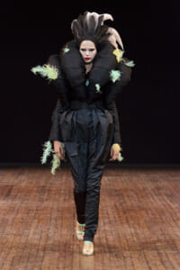 Runway model in an avant-garde black sculptural outfit with oversized collar, mint fringe details, and high-waisted pants; turquoise sandals visible.