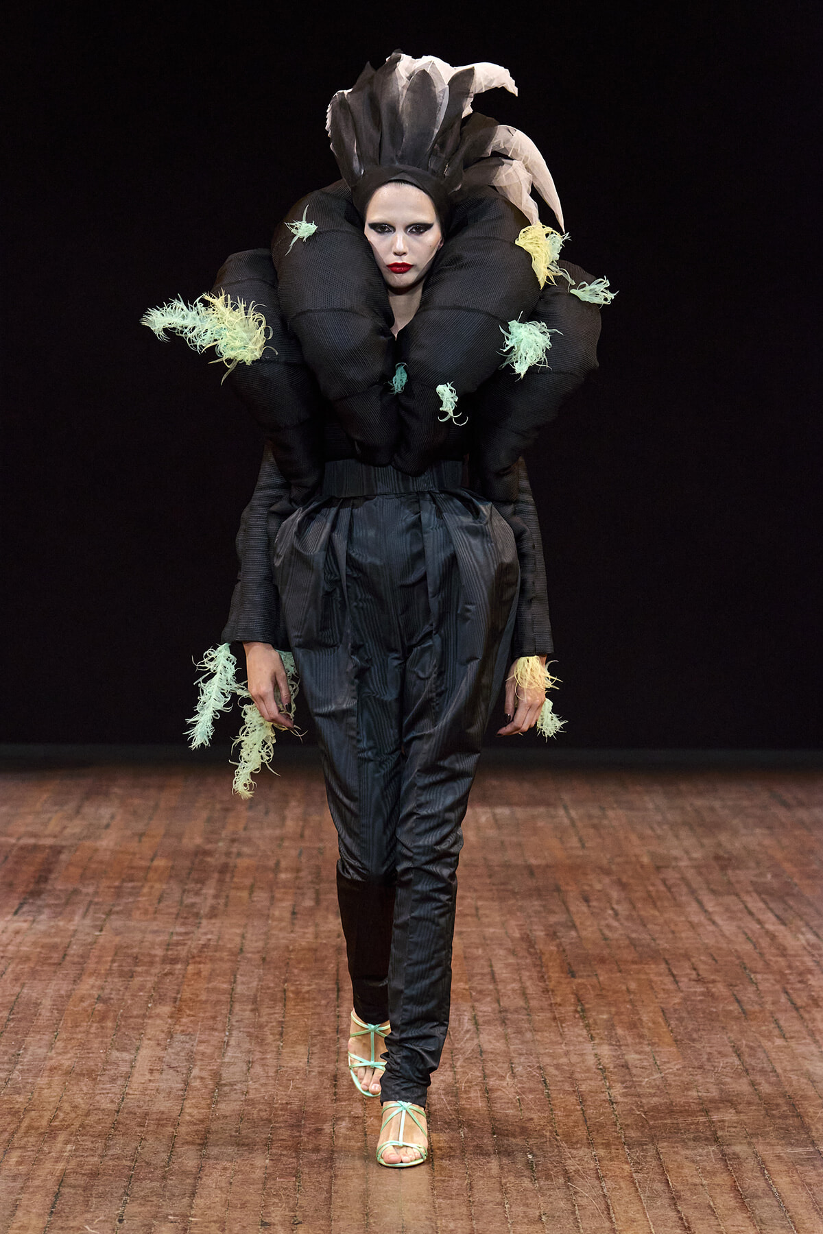 Runway model in an avant-garde black sculptural outfit with oversized collar, mint fringe details, and high-waisted pants; turquoise sandals visible.