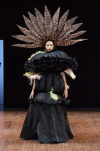 Model on a runway in a dramatic black gown with oversized ruffles and a fan-like feather headdress.