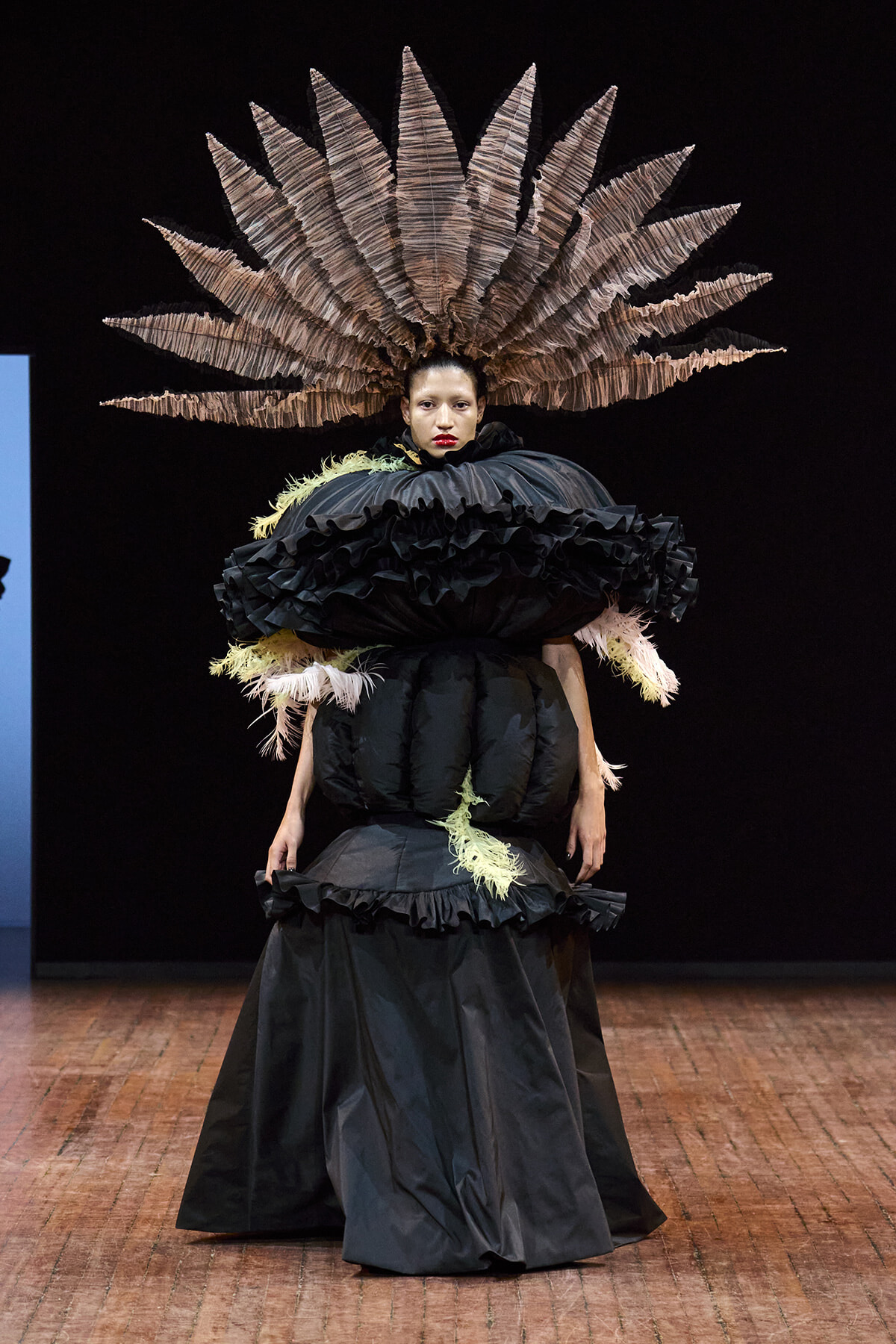 Model on a runway in a dramatic black gown with oversized ruffles and a fan-like feather headdress.