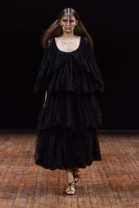 Runway model in a voluminous black textured, tiered dress with strappy sandals on a dark catwalk.