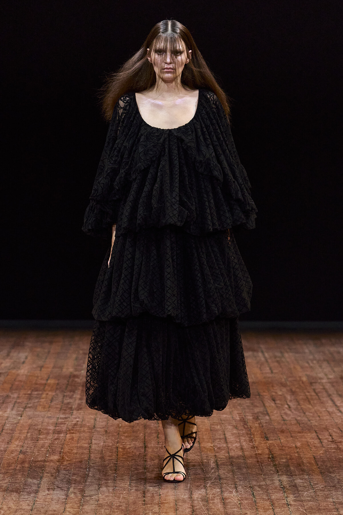 Runway model in a voluminous black textured, tiered dress with strappy sandals on a dark catwalk.