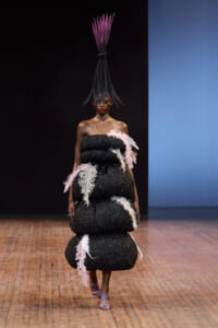 Model on a runway wearing a dramatic black glitter gown with pink and white feather accents and a tall purple feather headpiece.