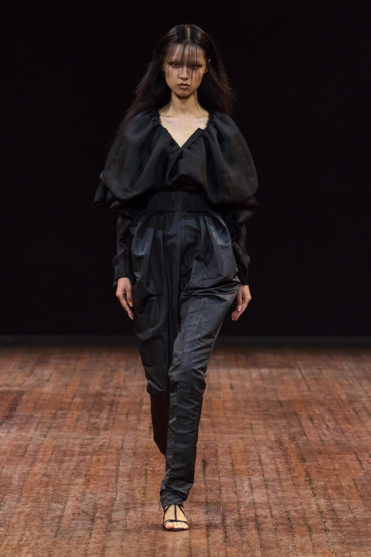 Model walking a fashion runway in an all-black, voluminous blouse with puffed sleeves and matching tapered pants on a wooden stage.
