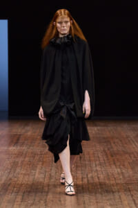 Model on a runway wearing a black, voluminous dress with a ruffled high collar, dramatic sleeves, and a large bow at the waist; red-haired model walking forward.