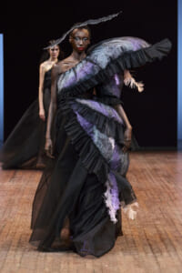 Model walks the runway in a dramatic black gown with purple lace, layered ruffles, and a large curved feather accessory.