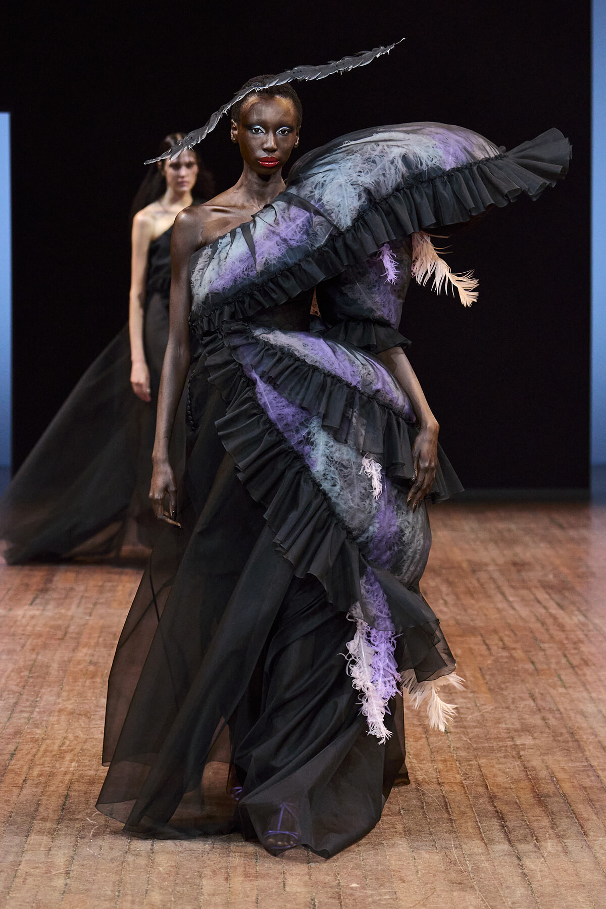 Model walks the runway in a dramatic black gown with purple lace, layered ruffles, and a large curved feather accessory.