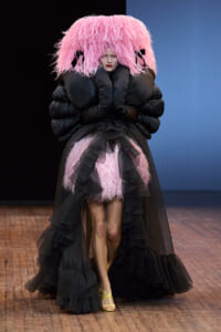 Model on runway in a dramatic black gown with a pink feather hood and oversized puffer sleeves.