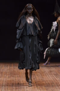 Model walking a fashion runway in a voluminous black satin gown with oversized puff sleeves and a deep neckline.