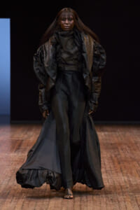 Runway model in an all-black, textured cape over a tailored black top and wide-leg pants, walking on a wooden floor runway.