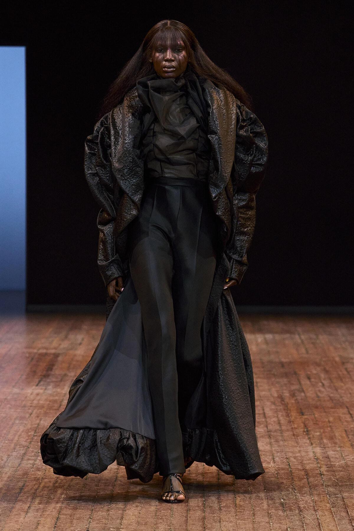 Runway model in an all-black, textured cape over a tailored black top and wide-leg pants, walking on a wooden floor runway.