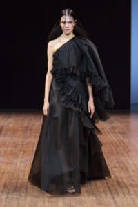 Model walks a fashion show in a black one-shoulder gown with cascading ruffles and a voluminous layered train on a wooden runway.