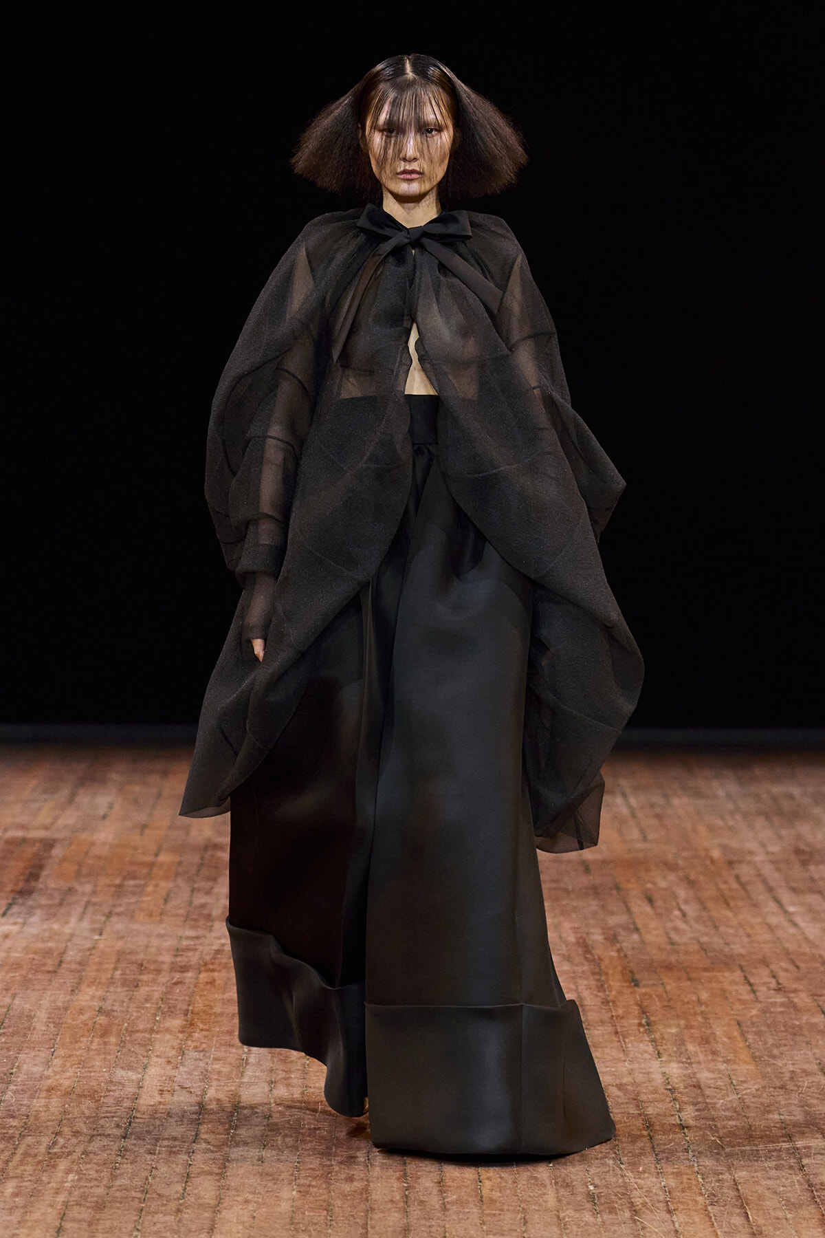 Runway model in a black, sculptural cape with sheer layers over a floor-length skirt, walking a fashion show stage.
