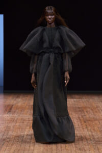 Model wearing a black couture gown with oversized cape-like sleeves on a runway, cinched at the waist with a belt.