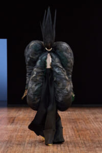 Runway model wearing a dramatic black sculptural coat with oversized puff sleeves and a tall vertical headpiece.