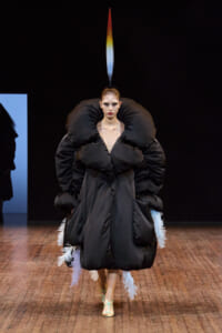 Model walking a fashion runway in an extreme black puffer dress with oversized voluminous sleeves and feather trim at the hem, topped by a tall rainbow-colored plume.