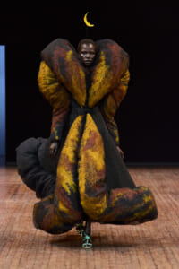 Fashion model on a runway wearing a towering, sculptural gown in yellow, orange, and black with feather-like textures; turquoise strappy heels visible.