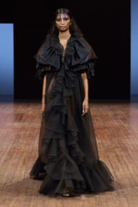 Model on a runway wearing a floor-length black gown with large layered ruffles and voluminous shoulders.