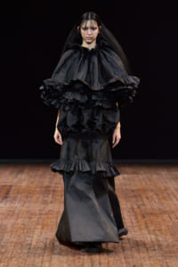 Model on a runway wearing a dramatic black ensemble with layered ruffles at the shoulders and a voluminous skirt.