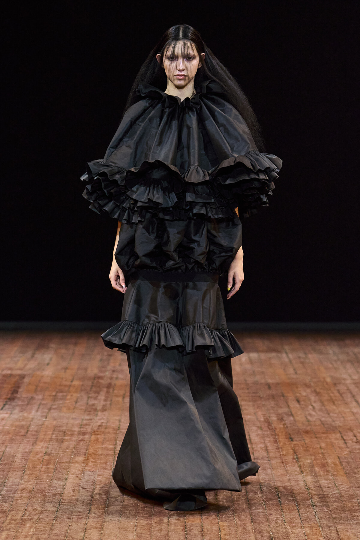 Model on a runway wearing a dramatic black ensemble with layered ruffles at the shoulders and a voluminous skirt.