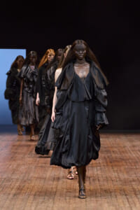 Model walks the runway in a dramatic, voluminous black gown with oversized ruffles; other models in dark outfits follow behind on a wooden runway.