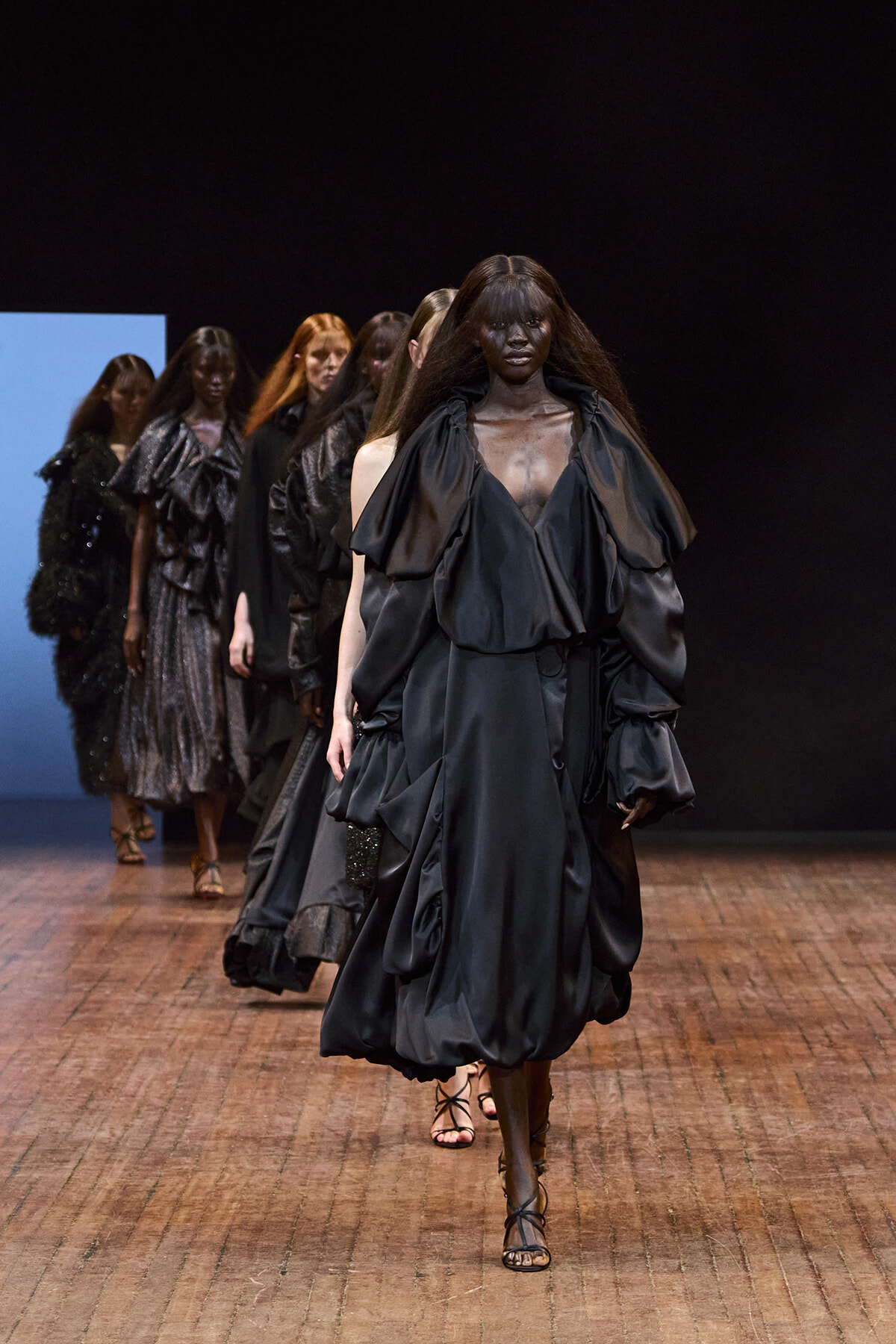 Model walks the runway in a dramatic, voluminous black gown with oversized ruffles; other models in dark outfits follow behind on a wooden runway.