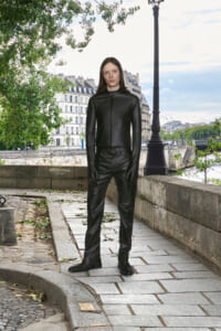 Person standing on a stone riverside path wearing a black leather jacket, gloves, and pants, with trees and a lamppost behind him/her.