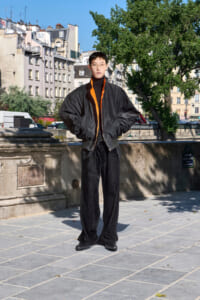 Young adult wearing an oversized black bomber jacket with an orange lining, standing with hands in pockets on a sunny urban promenade.