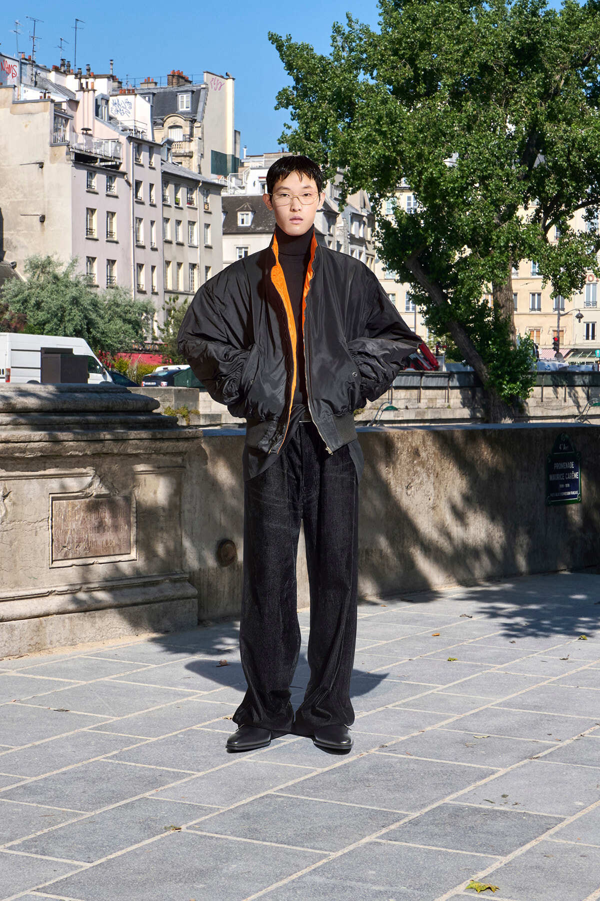 Young adult wearing an oversized black bomber jacket with an orange lining, standing with hands in pockets on a sunny urban promenade.