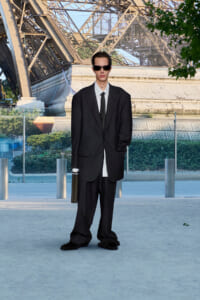 Man in an oversized black suit and sunglasses stands with a briefcase outdoors.